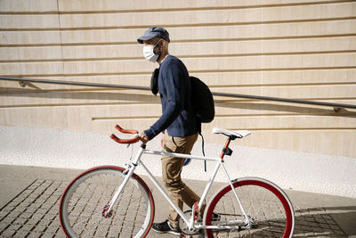 Man with bicycle looking away while walking by wall on footpath during covid-19