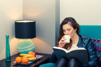 Woman reading book while drinking coffee on chair at home