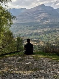 Rear view of man sitting on mountain against sky