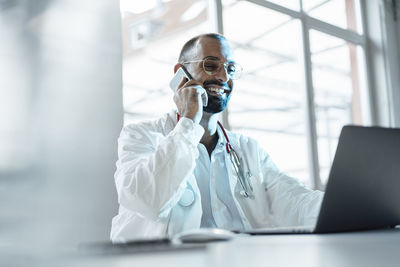 Happy doctor talking on mobile phone sitting with laptop on table in hospital