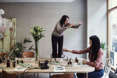 Young woman photographing while female colleague preparing perfume at table in workshop