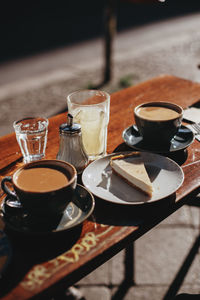 Cheese cake and two cups of coffee as well as fresh lemonade on a table