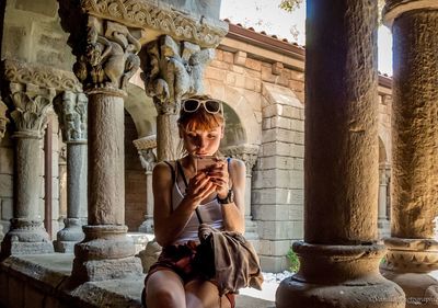 Woman using mobile phone while sitting on retaining wall at historic building corridor