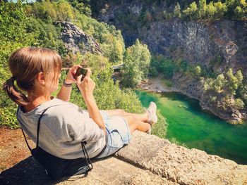 Woman photographing lake from cliff during sunny day
