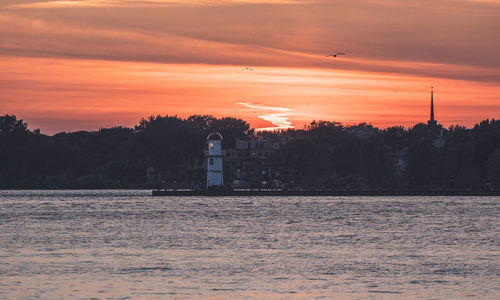 Silhouette of lighthosue by sea against orange sky