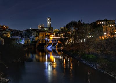 Illuminated bridge over river by buildings against sky at night