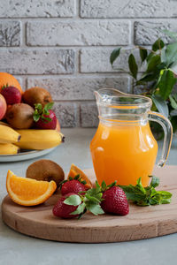 A jug of fruit juice, strawberry, orange and kiwi on a board. fruit on a platter in the background.