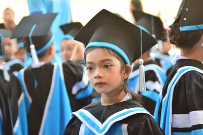 Close-up of girl wearing graduation gown and mortarboard looking away by friends