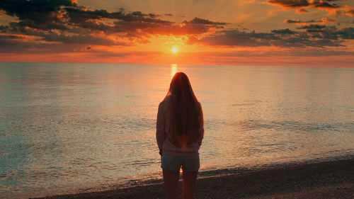 Rear view of woman standing at beach against sky during sunset