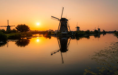 Traditional windmill by lake against sky during sunset