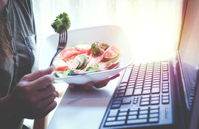 Midsection of woman working with salad bowl standing by laptop