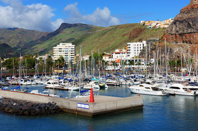 Sailboats moored at harbor by mountains against blue sky