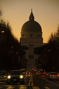 Cars on road by buildings against sky during sunset