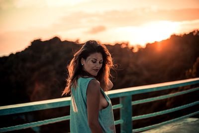 Young woman standing on footbridge against sky during sunset