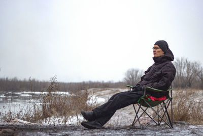 Brooding man sit on camping chair by frosty river, cold cloudy overcast weather with first snow