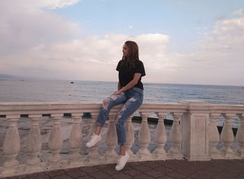 Young woman sitting on railing at promenade against cloudy sky during sunset