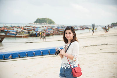 Portrait of smiling young woman holding camera while standing at beach