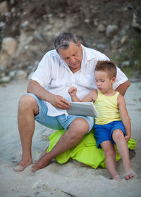 Grandfather with grandson using digital tablet while sitting at beach