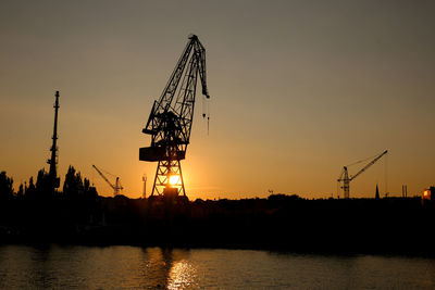 Silhouette cranes by river against sky during sunset