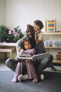 Male teacher reading book to preschool kids while sitting cross-legged in classroom at kindergarten