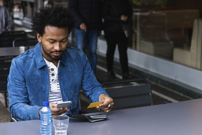 Mid adult man using credit card and smart phone for paying at sidewalk cafe