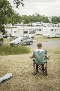 Rear view of boy sitting on camping chair while looking at travel trailers