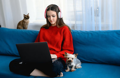 A young girl in a red  sweater works on a laptop at home. her cat and dog are sitting next to her