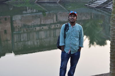 Young man with hands in pockets looking up while standing against river