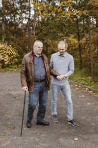 Full length of young male caretaker supporting retired senior man walking with cane on road
