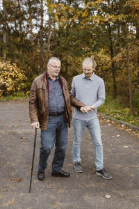 Full length of young male caretaker supporting retired senior man walking with cane on road