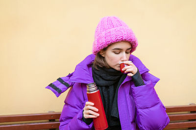 Young cute girl with dark hair in pink down jacket sits on wooden bench