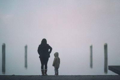 Full length of mother and child wearing warm clothing while standing by lake in winter during foggy weather
