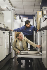 Mature customer looking at oven by saleswoman while crouching in electronics store