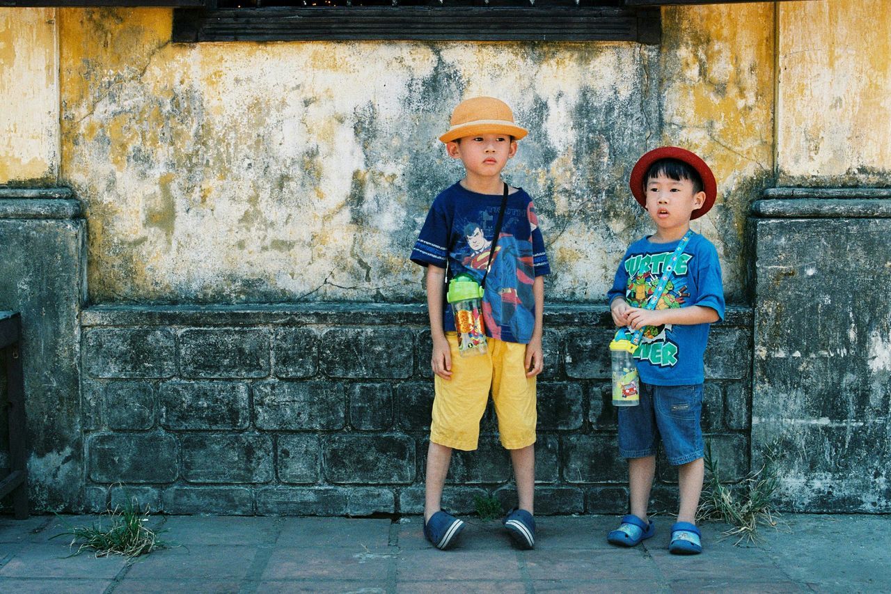 casual clothing, lifestyles, person, full length, front view, standing, wall - building feature, leisure activity, young adult, built structure, architecture, looking at camera, smiling, portrait, hands in pockets, building exterior, childhood