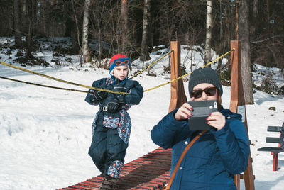 Boy looking at mother photographing while standing on field during winter