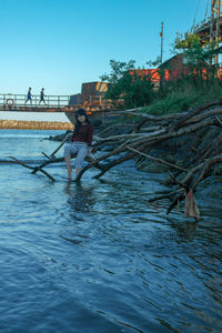 Teenage girl sitting on branch in river against clear sky