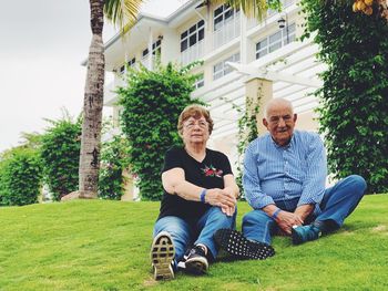 Full length of senior couple sitting on grassy field against building