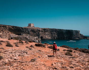 Rear view of woman with backpack walking on cliff against clear blue sky