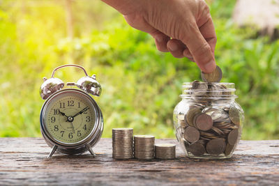 Cropped hand putting coins in jar by alarm clock on table