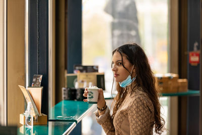 A beautiful teenage girl drinking coffee in bar wearing medical mask