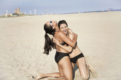 Happy female friends embracing while kneeling on sand at beach