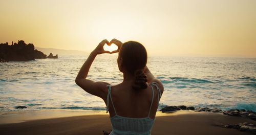 Rear view of woman with arms raised standing at beach against sky during sunset