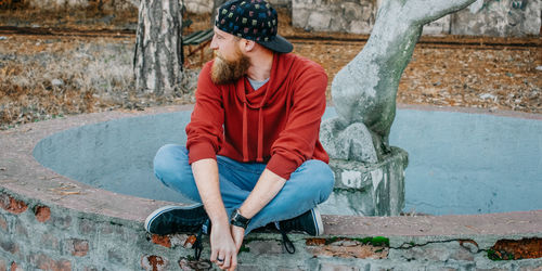 Young man looking away while sitting on retaining wall of fountain