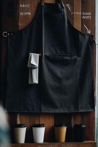 Close-up of black apron and disposable glass against wooden wall