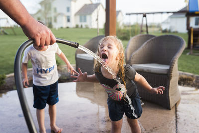 Cropped hand of man holding hose while girl drinking water and brother standing beside her at yard