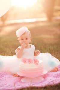 Baby girl 1 year old eating creamy birthday cake sitting on green grass with pink balloons outdoor