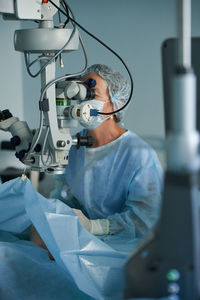 Focused adult female doctor in sterile mask and ornamental medical cap looking through surgical microscope against crop coworker in hospital