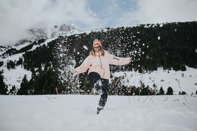 Young happy tourist in eyeglasses with upped leg looking at camera and having fun between field in snow near mountain