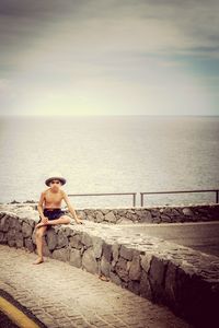 Shirtless boy looking away while sitting on retaining stone wall against sea