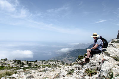 Side view of mature man with backpack sitting on mountain against sky